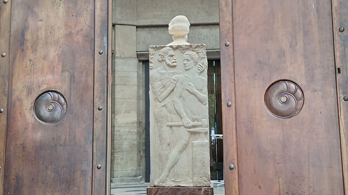 Blick durch die geöffnete Bronzetür des Abbe-Pavillons auf die Rückseite der Herme von Max Klinger: Das Flachrelief zeigt einen nackten Jüngling am Amboss stehend. Von hinten tritt Abbe, bekleidet mit einem dünnen weichen Gewand, heran und legt eine Hand auf den jungen Arbeiter.